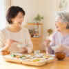 一人暮らしの高齢の母親と娘が、自宅のダイニングで宅配食を一緒に食べながら笑顔で話している様子
